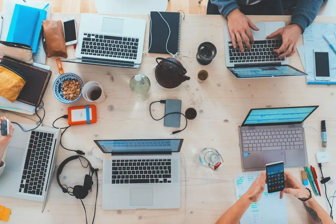 Overhead view of a busy shared workspace with multiple laptops, smartphones and IT equipment on a timber desk